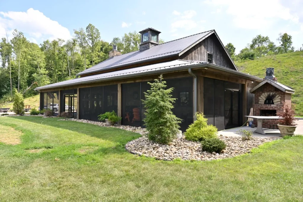 Exterior of a custom timber frame party barn with screened-in porch, metal roof, and brick outdoor fireplace. Surrounded by landscaped stone beds and greenery, this Sugarcreek Builder project highlights rustic barn design blended with modern outdoor living spaces.