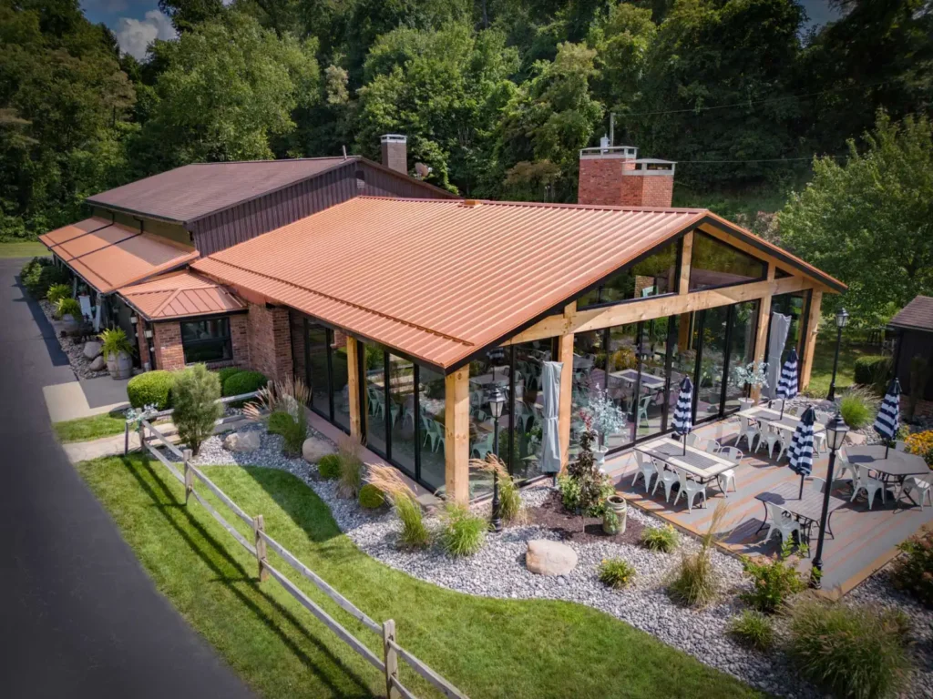 Exterior view of a custom timber frame lodge with glass walls, exposed beams, and a striking red metal roof. The design includes an outdoor patio with tables and umbrellas, landscaped stone beds, and a natural wooded backdrop—showcasing Sugarcreek Builder’s expertise in blending rustic craftsmanship with modern lodge architecture.