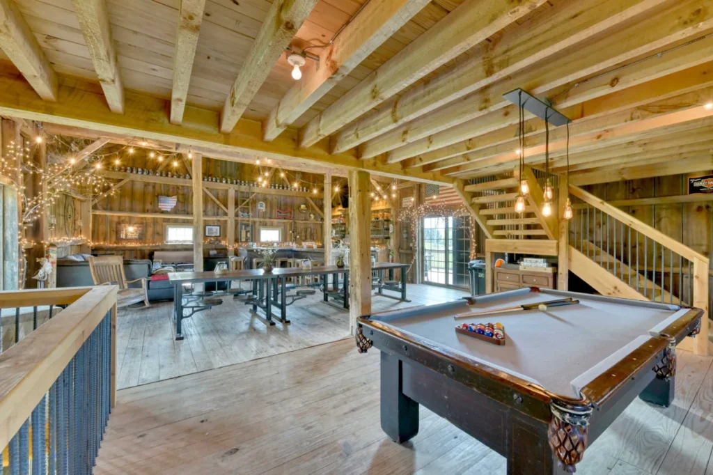 Rustic timber frame barn interior with exposed beams, wood ceiling, and entertainment area featuring a pool table, string lights, and custom seating—showcasing Sugarcreek Builder’s craftsmanship in recreational barn design.