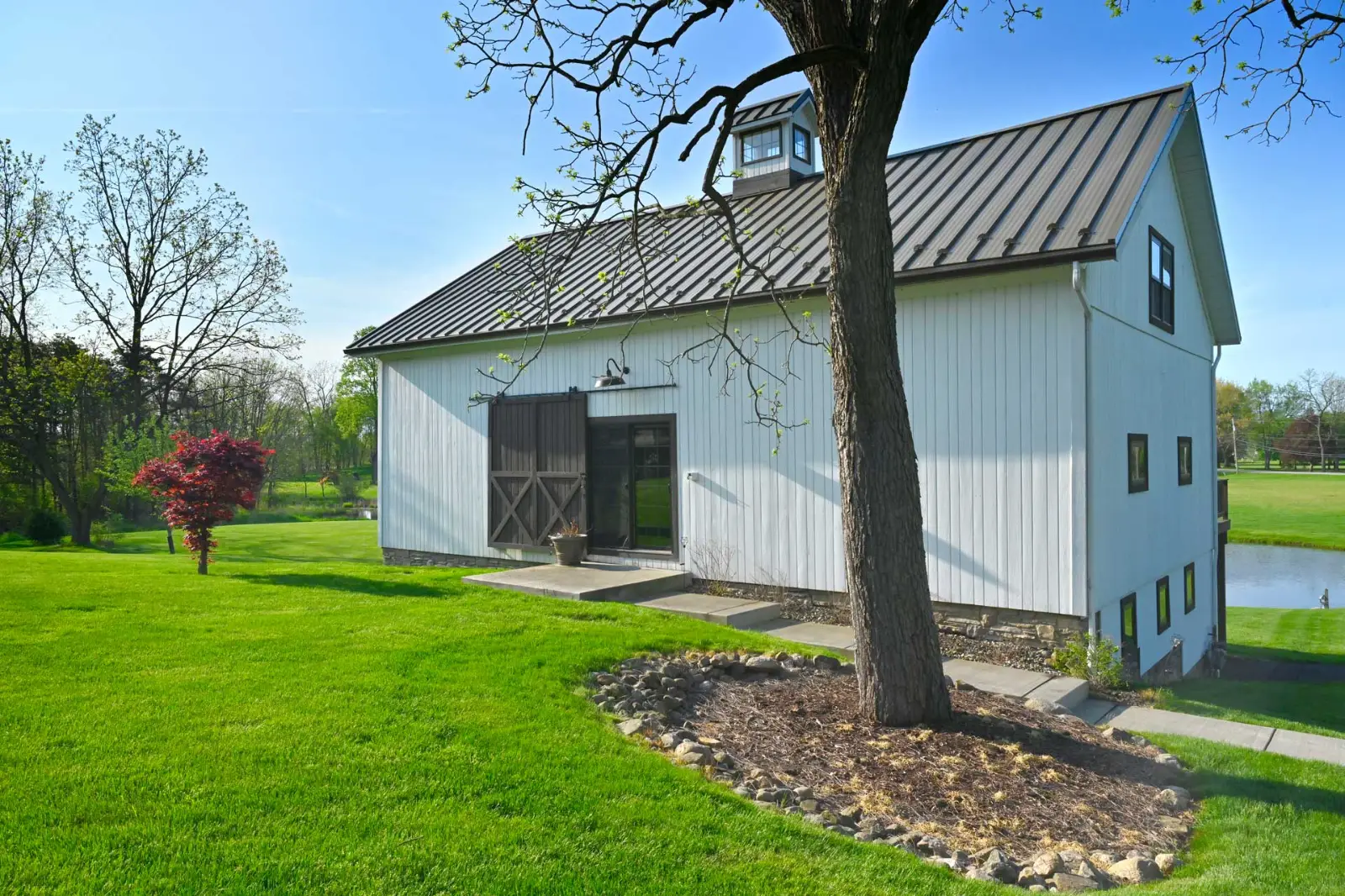 Exterior of Richardson Barn, a modern white barn with black metal roof, cupola, and sliding barn doors. Surrounded by open green lawn and mature trees, this Sugarcreek Builder project highlights traditional post-and-beam style with updated finishes.