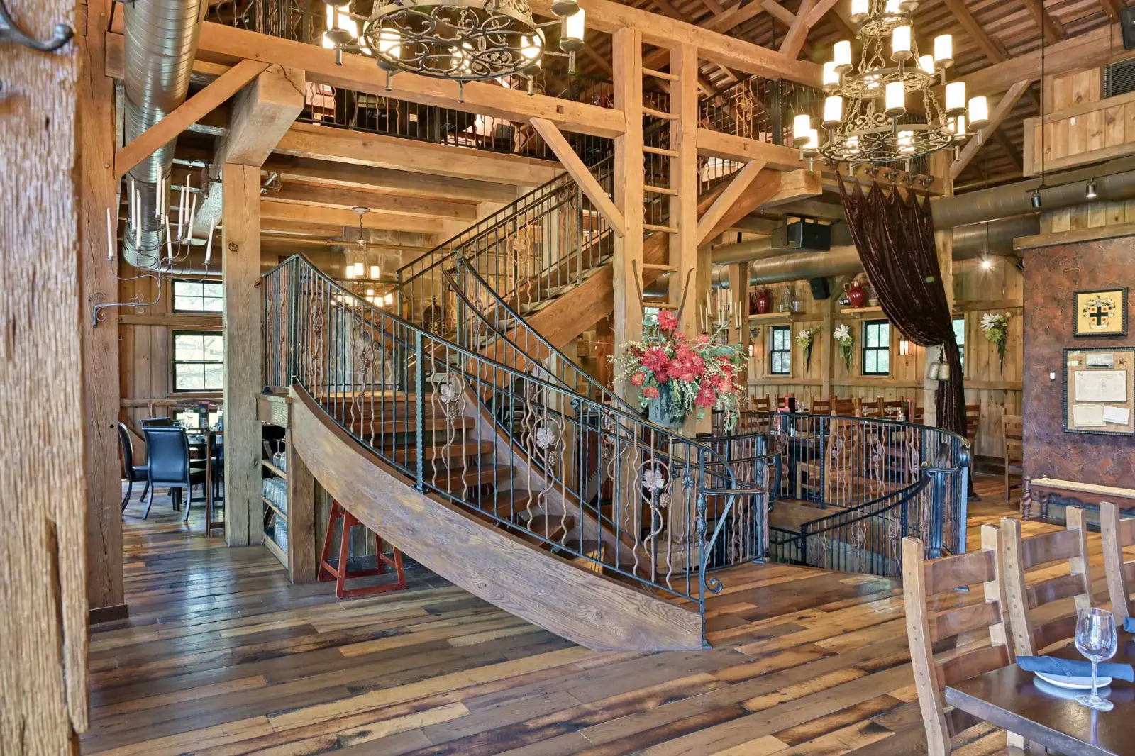 Interior of Gervasi Vineyard restaurant featuring timber frame beams, wrought iron staircase, chandeliers, and wood flooring. Sugarcreek Builder craftsmanship enhances the warm, rustic dining atmosphere.