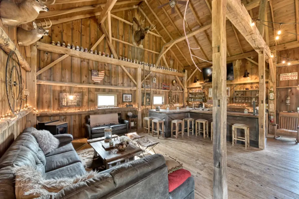 Rustic barn-style bar and lounge with exposed timber beams, wood-paneled walls, and vaulted ceiling. Features include a wraparound bar with wooden stools, leather sofas, mounted deer heads, and warm string lighting—showcasing Sugarcreek Builder’s custom barn interiors designed for entertaining.